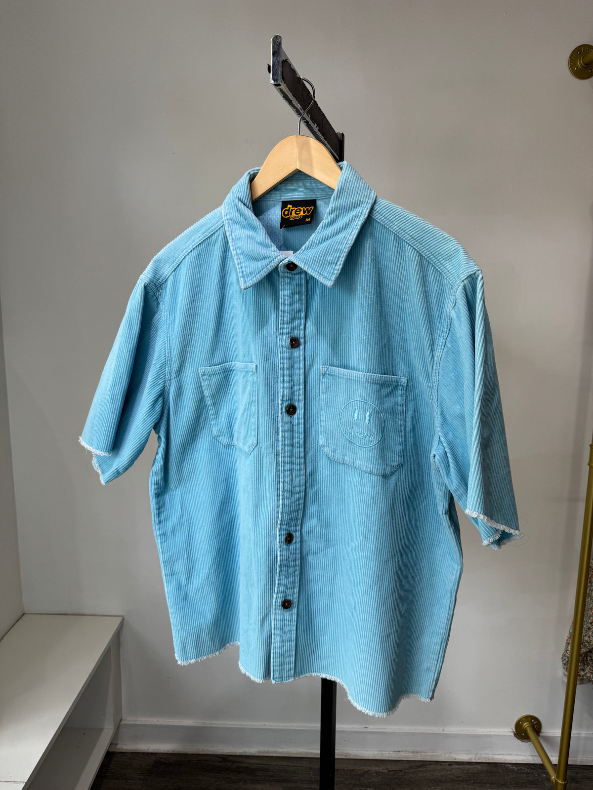A baby blue corduroy shirt on a hanger, displaying the front view with the brand name 'Drew House' visible on the collar.